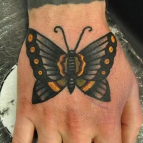 Mariposa en tonos oscuros, para el que se han utilizado algunos tonos de amarillo, verde y rojo que le dan un toque peculiar y distintivo a la mariposa tatuada sobre la mano, un gran tatuaje sin duda, que como podemos observar es el complemento a otro tatuaje que baja por el brazo y termina en la muñeca