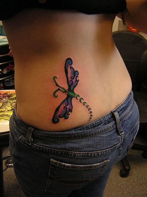 Tatuaje en la cintura de una libélula cuyas alas han sido construídas a partir de cenefas y un disimulado relleno en tonos lilas, por su parte, la libélula está trazada en tonos verdes y se ha hecho una original cola con trazos de medias circunferencias