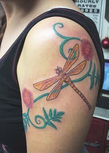 Tatuaje en el brao de una libélula en tonos amarillos y anaranjados, que ha sido completado con una espectacular cenefa de ramilletes de hojas en tonos turquesas y unas originales flores con los mismos colores que el cuerpo de la libélula