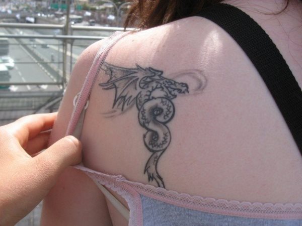 Tatuaje negro de dragón en el omoplato de esta chica, un tatuaje sencillo y simple que combina perfectamente con tonos de piel más clarito, ya que al no ser coloreado en negro al completo no resalta demasiado en una piel blanca