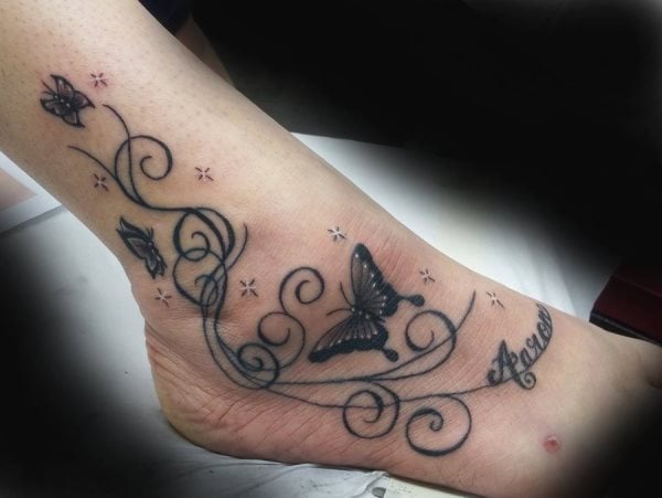 Tatuaje sobre el pie y el tobilo de unas pequeñas mariposas y trazos curvos para el que se ha tatuado en la parte final el nombre de alguien, una buena elección si es un tatuaje en honor de alguna persona