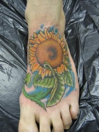 Tatuaje de un girasol sobre el pie que nos gusta mucho porque parece que el girasol tiene forma más humana que vegetal, gracias a la forma que se le han dado a las hojas y cuyos colores y fondo azul hacen de este un gran tatuaje