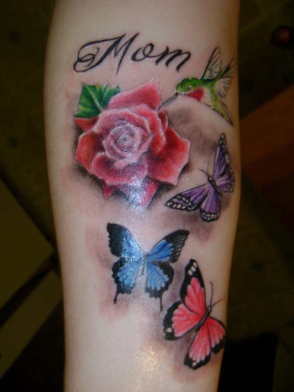 Tatuaje muy realista de rosas rojas y hijas verdes, custodiado por unas mariposas de varios colores y un pájaro verde de gran pico que llega volando para posarse sobre la flor, en este tatuaje se ha realizado un gran trabajo en la palabra mom por el toque a color blanco que se le ha dado a los trazos en negro