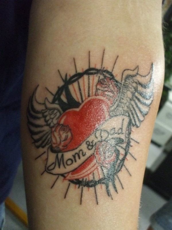 Tatuje de un corazón rojo con las palabras papá y mamá, para el cual se ha decidido por unas alas sobre el corazón y rodeado por una corona de espinas, en cuyo interior se han trazado unas líneas rectas en color negro y rojo que completan un tatuaje que inicialmente podría ser muy típico, pero que finalmente se ha conseguido un trabajo muy original