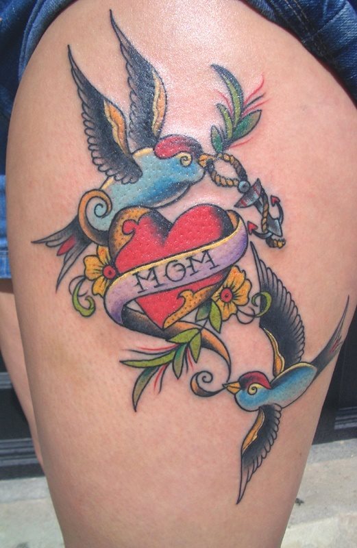 Completo y colorido tatuaje de un corazón rojo enlazado por la palabra mom y en cuyo corazón se ha posado un muy bien conseguido pájaro en colores que nos gustan muchísimo yen cuyo pico lleva una cuerda con un pequeño ancla y es que el amor hacia una madre siempre tiene que estar anclado en nuestro corazón y llevado con un mimo y delicadeza como la que lleva este pájaro el ancla