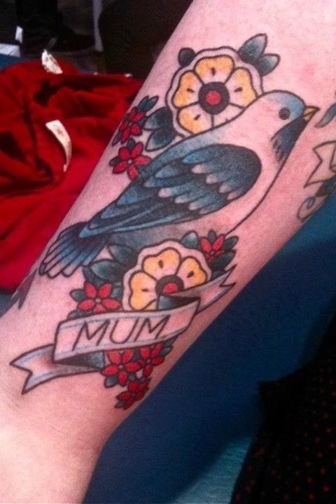 Tatuaje de una paloma en tonos negros y azules sobre unas flores blancas y amarillas, acompañadas de otras flores más pequeñas de color rojo